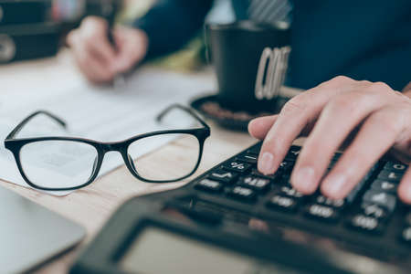 Cup of coffee and glasses on table and Accountant working on calculator to calculate company data, document and report on desk office. Accounting business concept.の写真素材