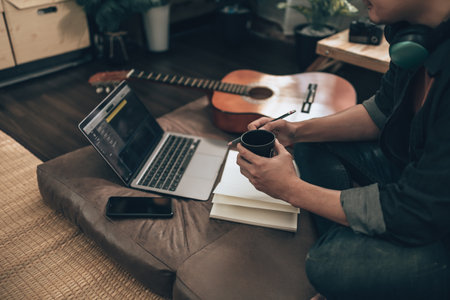 young man relax and playing guitar while sitting on sofa bed in living room at home. Music create melody song, lyrics on laptop and practice concept.の写真素材