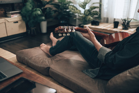 young man relax and playing guitar while sitting on sofa bed in living room at home. Music hobby and practice concept.の写真素材