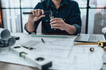 Engineering hands hold vernier caliper working and checking joint pipe scale with drawings inspection on the office desk and Calculator, triangle ruler, safety glasses, compass, vernier caliper on Blueprint. Engineer, Architect, Industry and factory concept.の写真素材