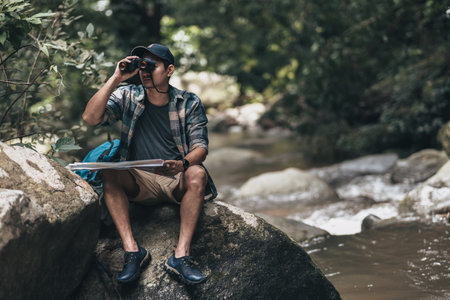 The trekker was looking at the map in his hand and use binocular to find the hiking trails at the stream in the forest. hiking and adventure concept.の写真素材