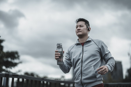 A man holding a water bottle and running up on footbridge in the city center park for cardio workout.  Health and Lifestyle in big city life concept.の写真素材