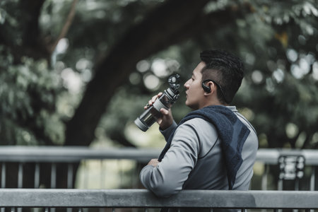 Young sport man listen music earphone and  drink water after jogging, running in the park. Sport thirsty and resting after exercise. Health and Lifestyle in big city life concept.の写真素材