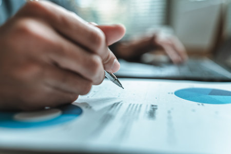 A close-up view of a businessman hand gripping a pen, indicating a project along with financial report on paper document, symbolizing data analysis, corporate, team and business strategy planning.の写真素材