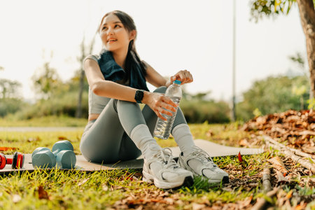 A woman dressed in workout clothes, feeling thirsty, is opening a water bottle for drink during a break from exercising in the park.の写真素材
