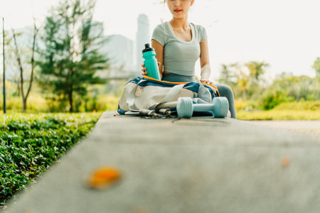 A woman in sportswear sits on a park bench with a gym bag, water bottle, dumbbells, and jump rope, preparing for an outdoor workout surrounded by nature and greenery.の写真素材
