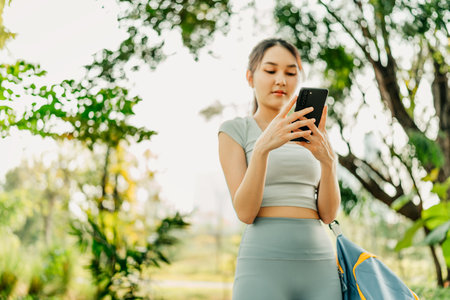 A woman in sportswear stands on a park path, holding a phone and gym bag, surrounded by lush greenery and trees, enjoying a peaceful, healthy outdoor environment.の写真素材