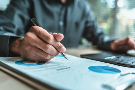 A close-up view of a businessman hand gripping a pen, indicating a project along with financial report on paper document, symbolizing data analysis, corporate, team and business strategy planning.の写真素材