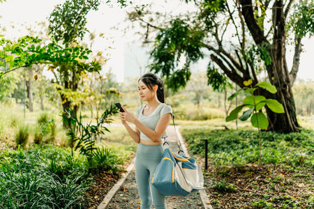 A woman in sportswear stands on a park path, holding a phone and gym bag, surrounded by lush greenery and trees, enjoying a peaceful, healthy outdoor environment.の写真素材