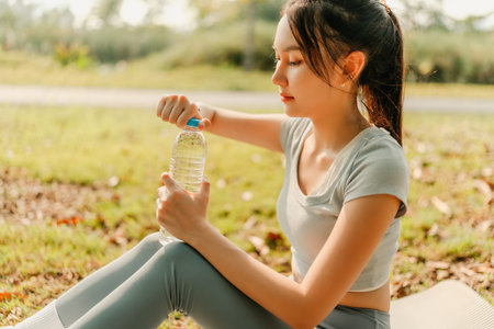 A woman dressed in workout clothes, feeling thirsty, is opening a water bottle for drink during a break from exercising in the park.の写真素材