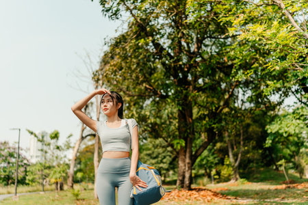 A woman is walking along a paved path in a green urban park, dressed in athletic wear including leggings, a crop top, and sneakers, and carrying a duffel bag.の写真素材
