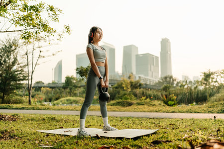 A woman dressed in workout clothes is wearing headphones, listening to music, and performing squats with kettlebell to exercise her legs and glutesの写真素材