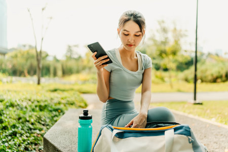 A woman in sportswear sits on a park bench with her phone, water bottle, and gym bag, enjoying a break in a green urban park on a sunny day.の写真素材