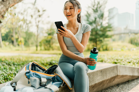 A woman in sportswear sits on a park bench with her phone, water bottle, and gym bag, enjoying a break in a green urban park on a sunny day.の写真素材