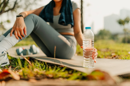 A woman dressed in workout clothes is reaching for a water bottle placed beside her during a break from exercising. Workout, Fitness, Sport and Yoga concept.の写真素材