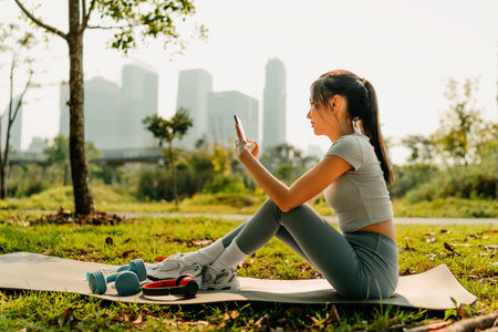 A woman dressed in workout clothes is sitting on a yoga mat, using her phone with blank screen. headphones, dumbbells and a water bottle on the mat. Exercise concept.の写真素材