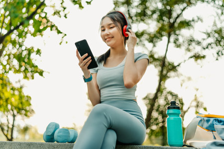 A woman dressed in workout clothes is using her phone and wearing headphones to listen to relaxing music before exercising at the park.の写真素材