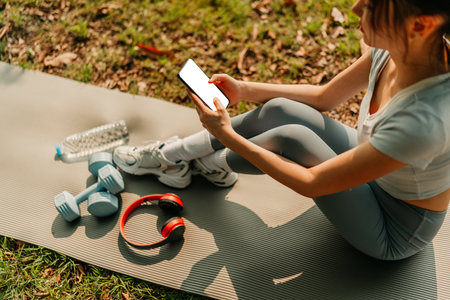 A woman dressed in workout clothes is sitting on a yoga mat, using her phone with blank screen. headphones, dumbbells and a water bottle on the mat. Exercise concept.の写真素材