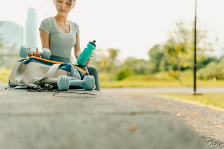 A woman in sportswear sits on a park bench with a gym bag, water bottle, dumbbells, and jump rope, preparing for an outdoor workout surrounded by nature and greenery.の写真素材