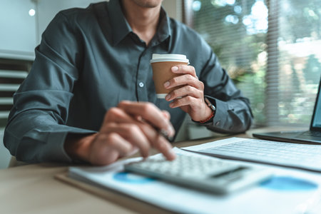 Businessman drink hot coffee. An accountant is seated at a desk in the office, utilizing a calculator to determine balances, profits, currency conversions, and costs.の写真素材