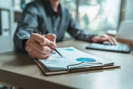 A close-up view of a businessman hand gripping a pen, indicating a project along with financial report on paper document, symbolizing data analysis, corporate, team and business strategy planning.の写真素材