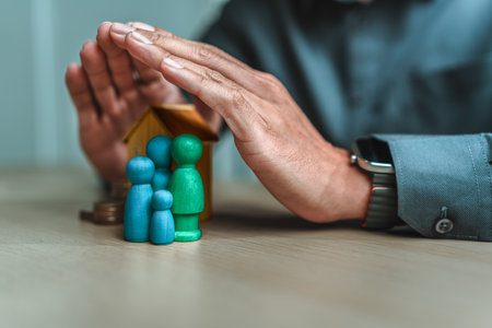 Businessman's hand gestures to protect money, house, wooden dolls represent family. Image represents the concept of asset protection, emphasizing safeguarding financial investments and propertyの写真素材