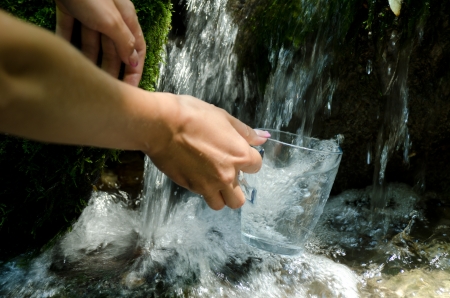 Glass of water by waterfall. Human hand holding glass pouring fresh drink water.の写真素材