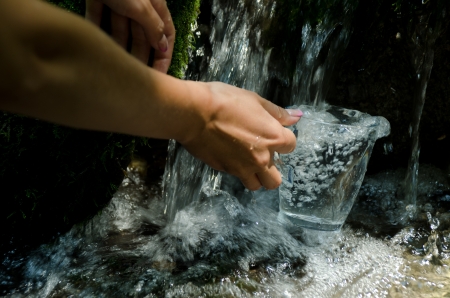 Glass of water by waterfall. Human hand holding glass pouring fresh drink water.の写真素材