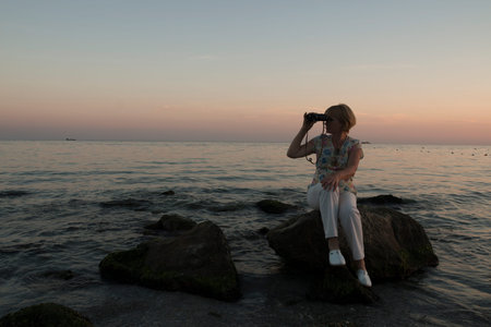 Woman sitting on the stones looking in binocular. Ocean background.の写真素材