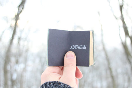 Adventure, hand holding a book with text on background of winter park