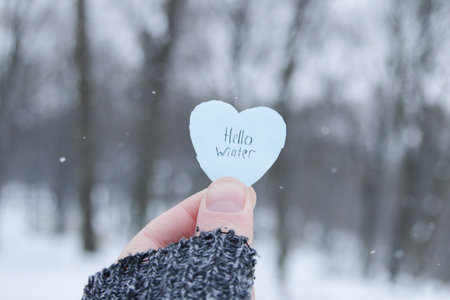 hello winter. A hand holds a paper blue heart with the inscription on the background of the winter forest.の写真素材