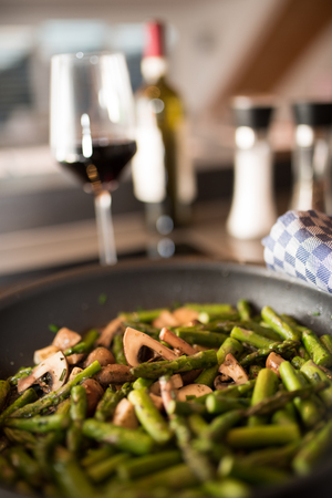 Fried green asparagus with mushrooms in a pan in front of a kitchen backgroundの写真素材