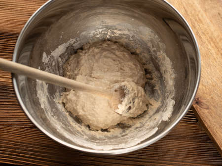 Dough in a metal bowl for a homemade baguette. Mixing of ingredients with a wooden spoon on a wooden kitchen counter. Top view photography.の写真素材