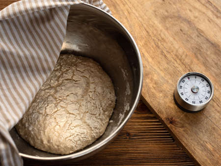 Fresh dough in a metal bowl for a homemade baguette on a wooden kitchen counter with alarm clock. Top view photography.の写真素材