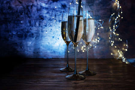 New year celebration with sparkeling champagne. Champagne glasses with drinking straw on wooden bar counter against luminous blue background with fairy lights.の写真素材