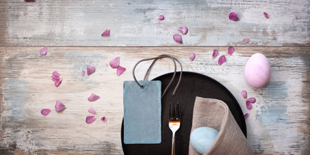 Easter dinner on gray vintage wood with pink petals. Place setting with pink and bright blue easter...