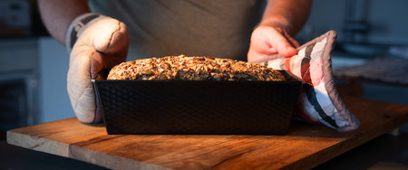 Hands hold freshly baked whole grain bread in hot baking pan on wooden work surface. Close-up.