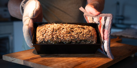 Hands hold freshly baked whole grain bread in hot baking pan on wooden work surface. Close-up.の写真素材