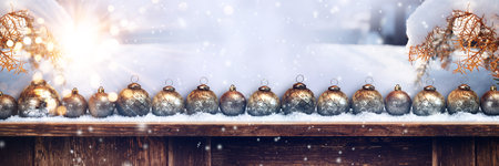 Christmas greeting card. Row of silver Christmas balls in the snow on an old wooden counter. Glistening rays of light and festive golden bokeh. Winter background with snowflakes and snowy branches.の写真素材
