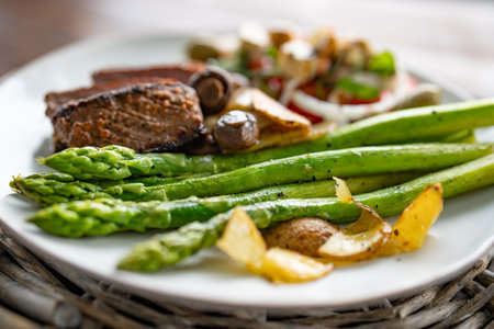 Roasted green asparagus with a beef steak, mushrooms, and crispy potato slices served on a white plate. Close-up with shallow depth of field.の写真素材