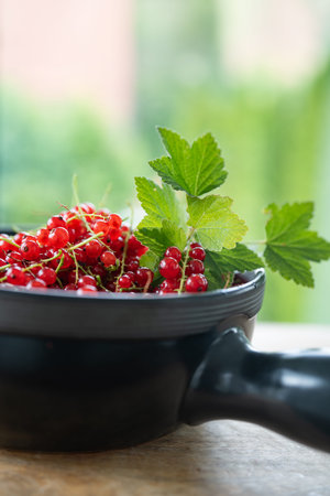 Freshly picked ripe red currants in a dark ceramic bowl, decorated with green leaf on wooden table...