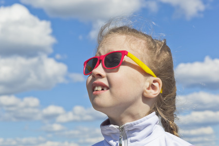 Dreamy small girl wear sunglasses against the sky with clouds looking at the topの写真素材