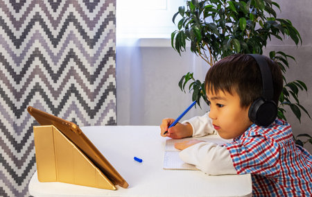 A smart young boy is sitting at a table, writing homework or preparing for an exam. Boy using laptop...