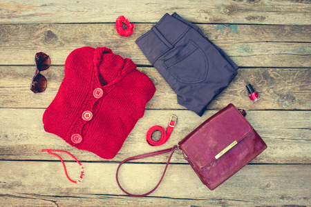 Women's autumn clothing and accessories: red sweater, pants, handbag, beads, sunglasses, nail polish, hair band, belt on wooden background. Toned image.の写真素材