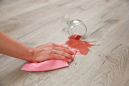 Glass of red wine fell on laminate, wine spilled on floor. A woman wipes the laminate from moisture.の写真素材
