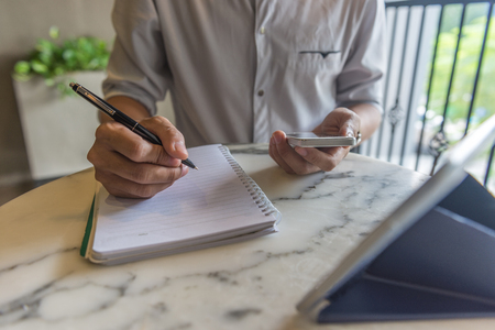 Young freelancer using smartphone and writing into noteの写真素材