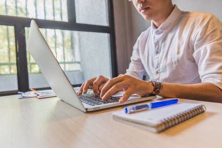 Asian man in white shirt typing laptop next to windowの写真素材