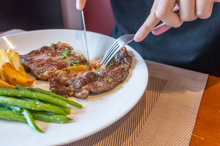 Using knife and fork cutting beefsteak on white plate