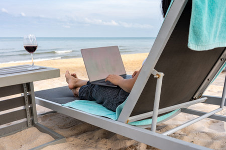 Rear view of woman working on laptop with glass of wine on exotic beach