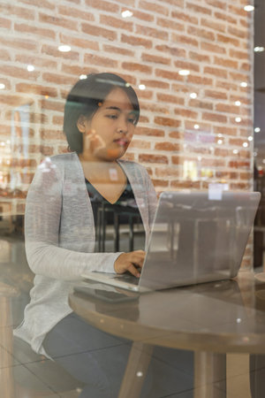 Through glass window view of woman using laptop at coffeeshop